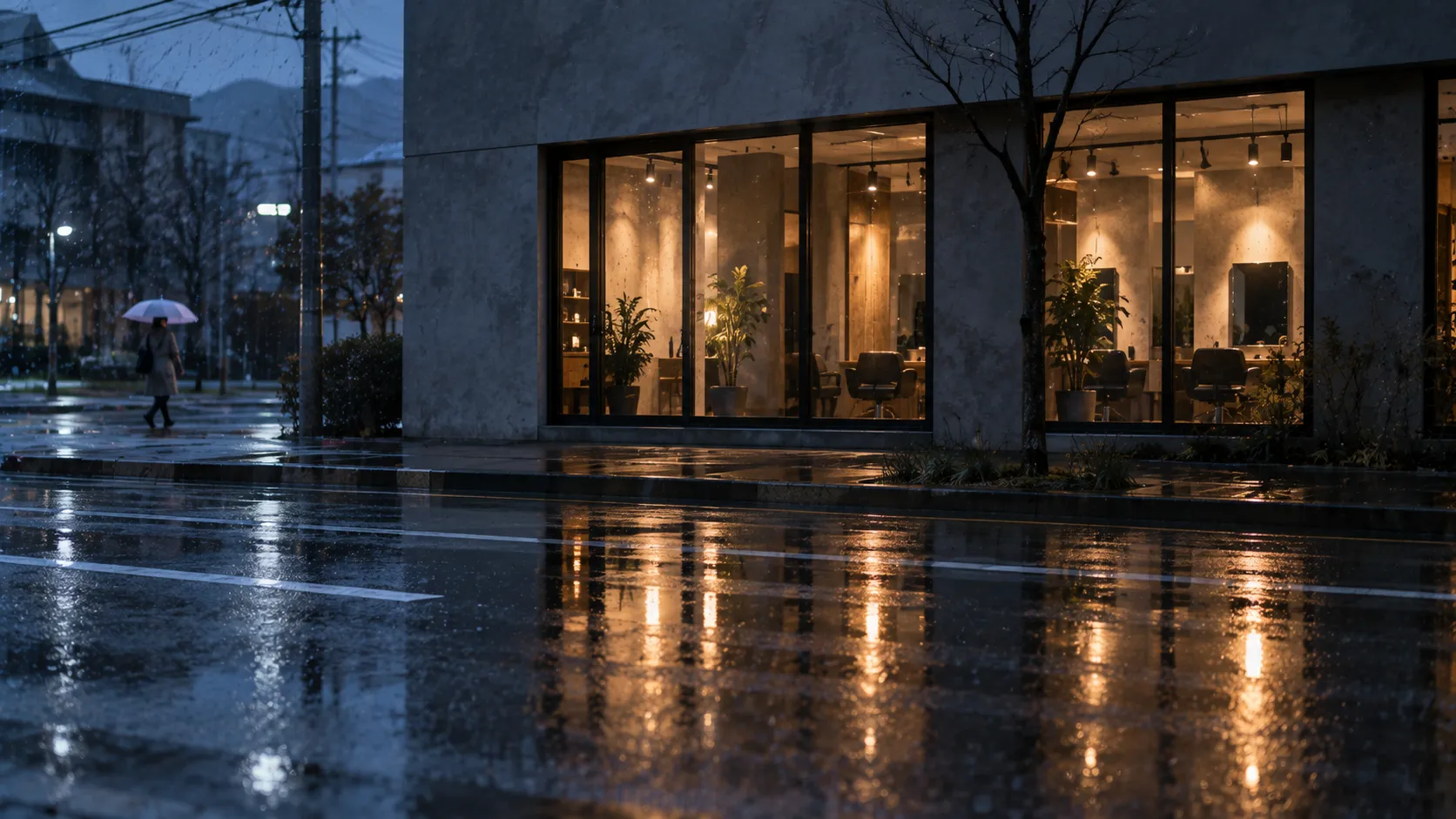 雨の日没後に濡れた路面へ店の窓の灯りが伸びる通りからの引き