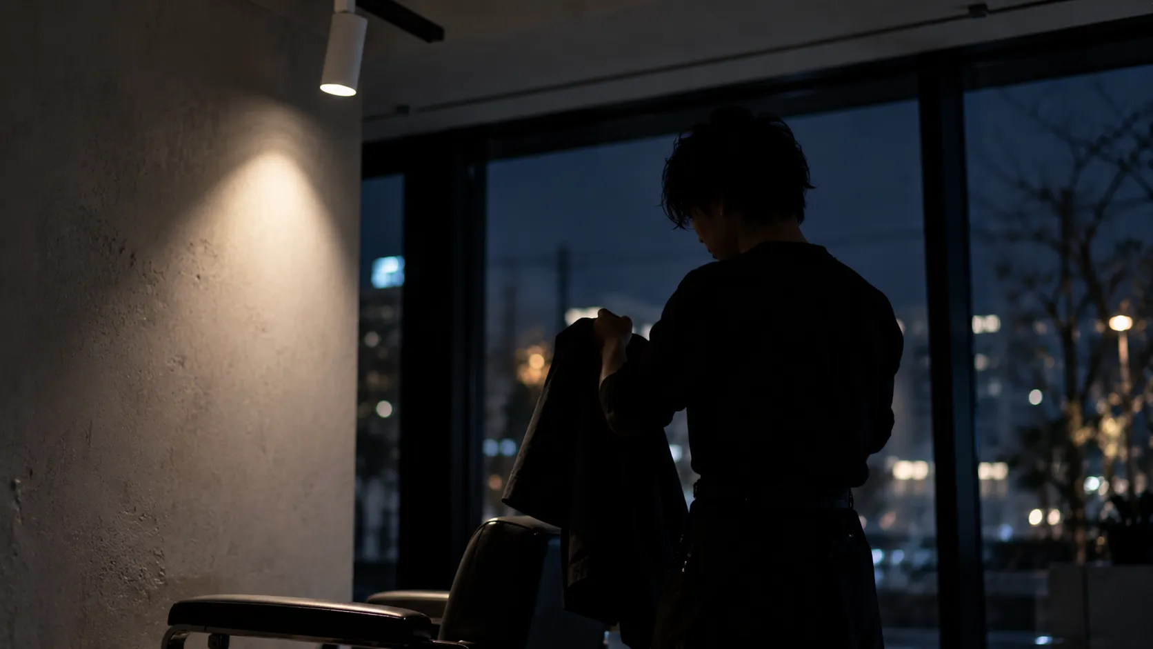 夜、店の灯りを背に帰り支度をする男性スタイリストのシルエット